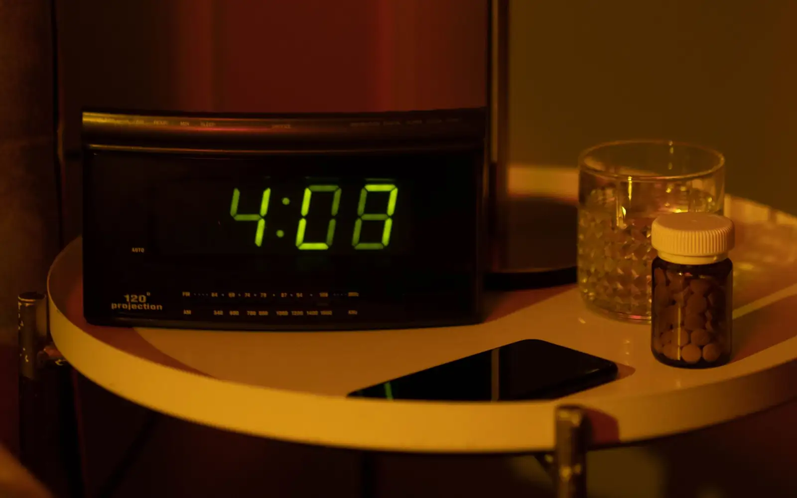 Despertador digital, um copo de água e um frasco de comprimidos numa mesa de cabeceira à noite